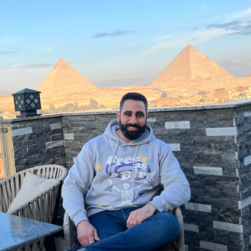 مجدي كميل، الخبير في الاقتصاد الرقمي، يجلس بثقة وخلفه أهرامات الجيزة؛ تجسيدًا لنهج بناء استراتيجيات القيمة والاستدامة الرقمية.